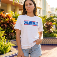 A woman wearing a white graphic t-shirt with 'CUBAN BRED' printed in blue and red, paired with denim shorts, standing outdoors in front of a sunny building with tropical plants.