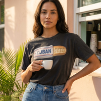 A woman wearing a black graphic t-shirt with a 'CUBAN BRED' design, holding a white coffee mug in front of a warm-toned building wall with palm leaves – Cuban American graphic tee, Miami gift by Martha of Miami