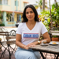A woman sitting at an outdoor cafe table wearing a white v-neck t-shirt with 'CUBAN BRED' printed in blue and red across the chest, paired with light blue jeans.