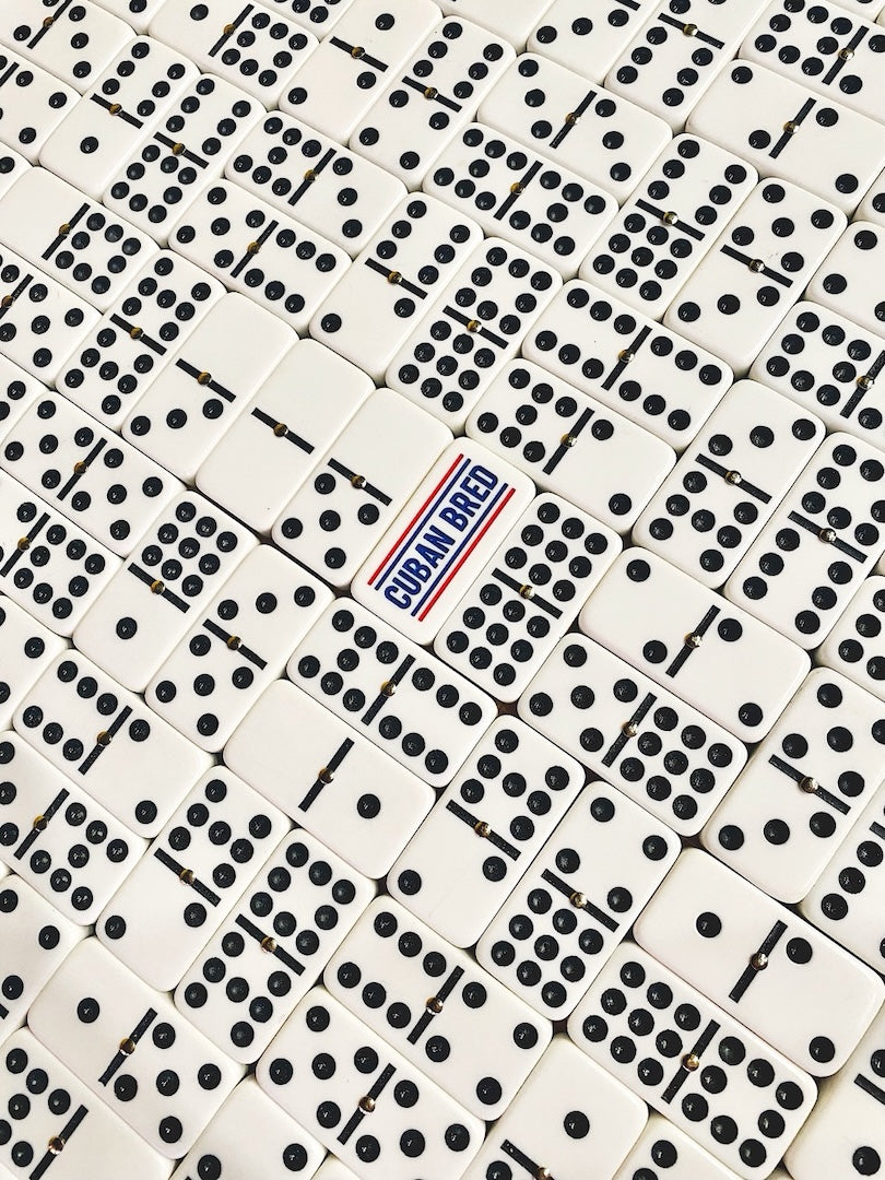 A close-up overhead view of many white domino tiles with black pips, arranged in a grid. One central domino is different, with the words "Cuban Bred" printed in blue and red.
