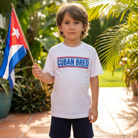 A young boy stands outdoors on a tiled patio holding a Cuban flag, wearing a white short-sleeve t-shirt with 'CUBAN BRED' printed across the chest in blue with red stripes, and dark navy shorts.