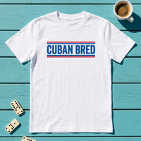 A white short-sleeve t-shirt with 'CUBAN BRED' printed in blue block letters between two red horizontal stripes, laid flat on a turquoise wooden surface with a coffee cup, dominoes, and a small plant nearby.