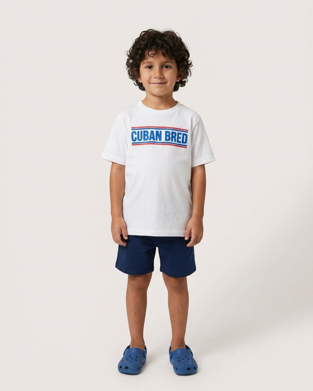 A young boy with curly dark hair stands against a light gray studio background wearing a white short-sleeve T-shirt with a blue and red &