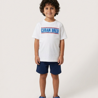 A young boy with curly dark hair stands against a light gray studio background wearing a white short-sleeve T-shirt with a blue and red 'CUBAN BRED' graphic across the chest, navy shorts, and blue clogs.