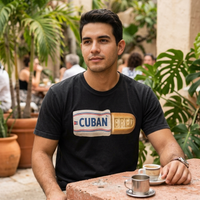 A man sitting at a stone table in a tropical patio setting, wearing a black t-shirt with a 'CUBAN BRED' graphic in a Cuban flag-inspired design, a small coffee cup on the table – Cuban American graphic tee, Miami gift by Martha of Miami