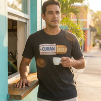 A man wearing a black graphic t-shirt with a 'CUBAN BRED' design, standing outdoors next to a turquoise building and holding a white coffee cup – Cuban American graphic tee, Miami gift by Martha of Miami