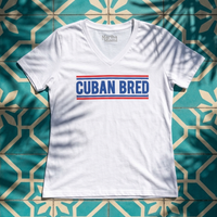 A white short-sleeve V-neck t-shirt with 'CUBAN BRED' printed in blue block letters between two red stripes across the chest. The shirt is laid flat on a teal and beige geometric tile background with dappled shadows.