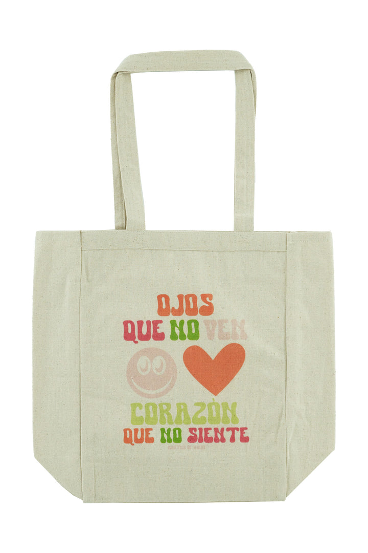 an image of a canvas tote bag with color text that reads in spanish Ojos que no ven, corazon que no siente with a smiley face and a heart graphic
